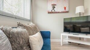 A modern living room with a blue sofa, a fluffy grey throw, and a flat-screen TV on a white stand.