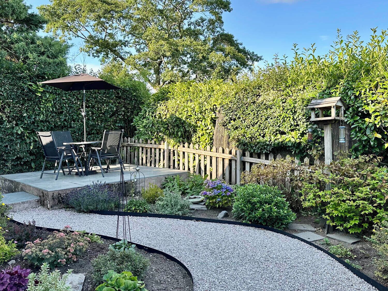 Une terrasse de jardin avec une table et des chaises, entourée d’une clôture en bois et de diverses plantes.