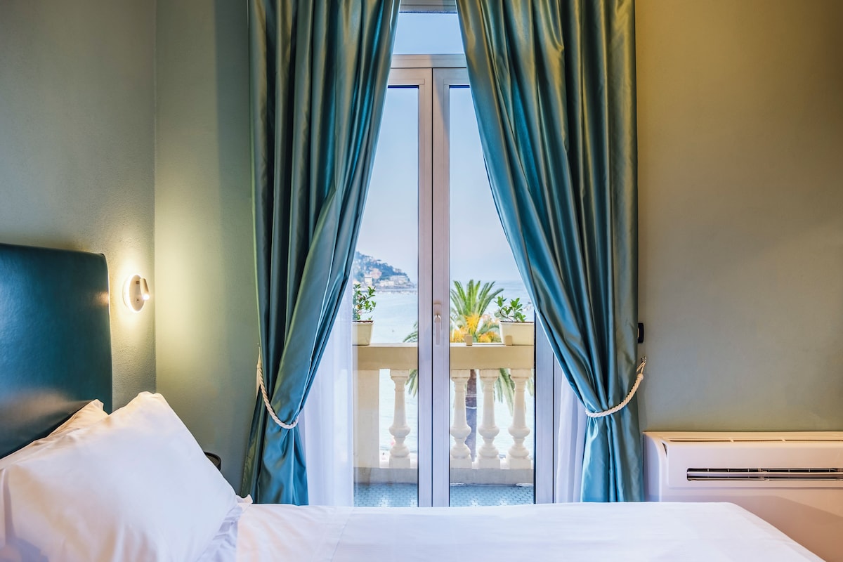 A hotel room with a bed, curtains, a view of the sea, and a balcony with potted plants.