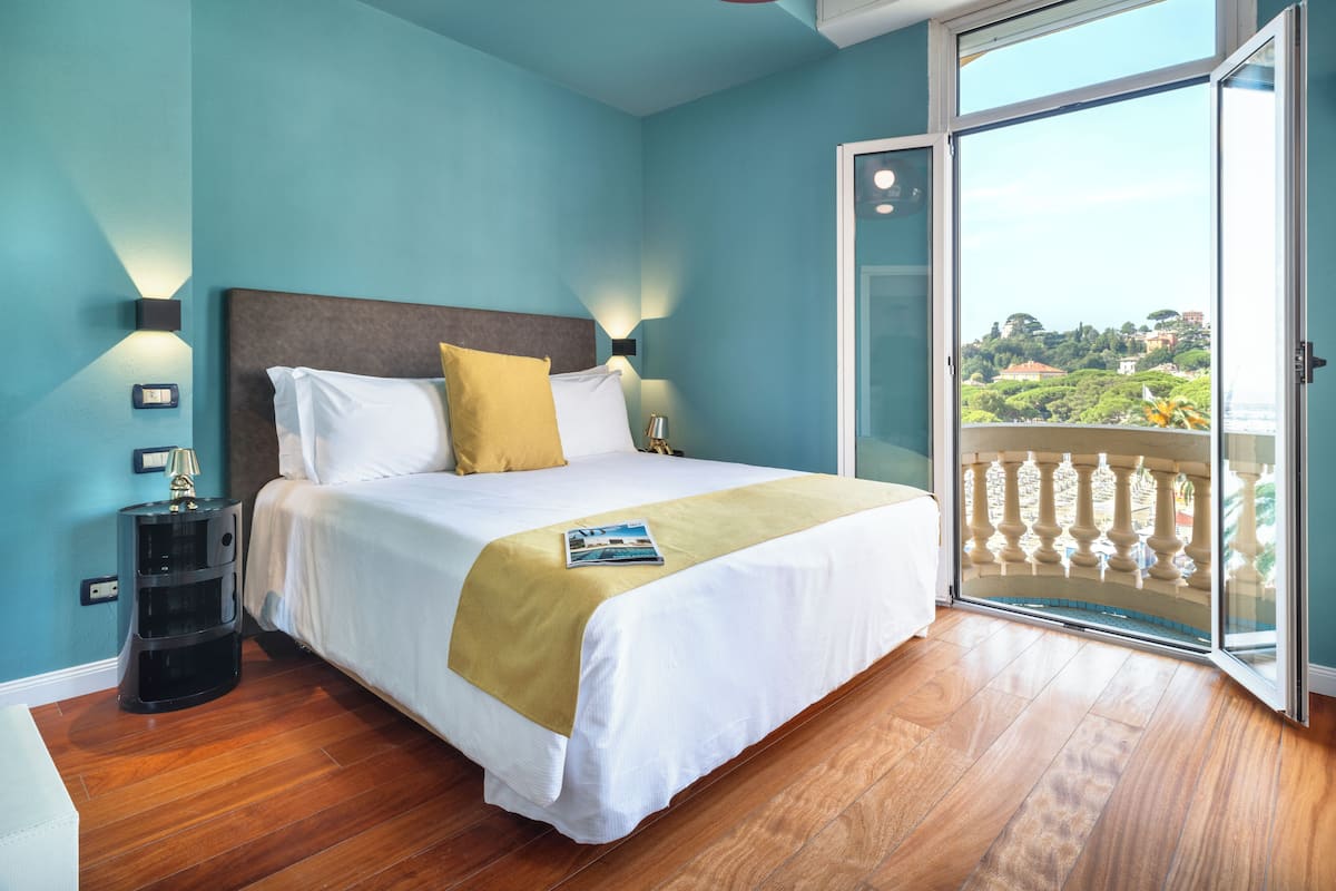 A hotel room with a bed, a nightstand, a lamp, and a view of the city through a balcony.