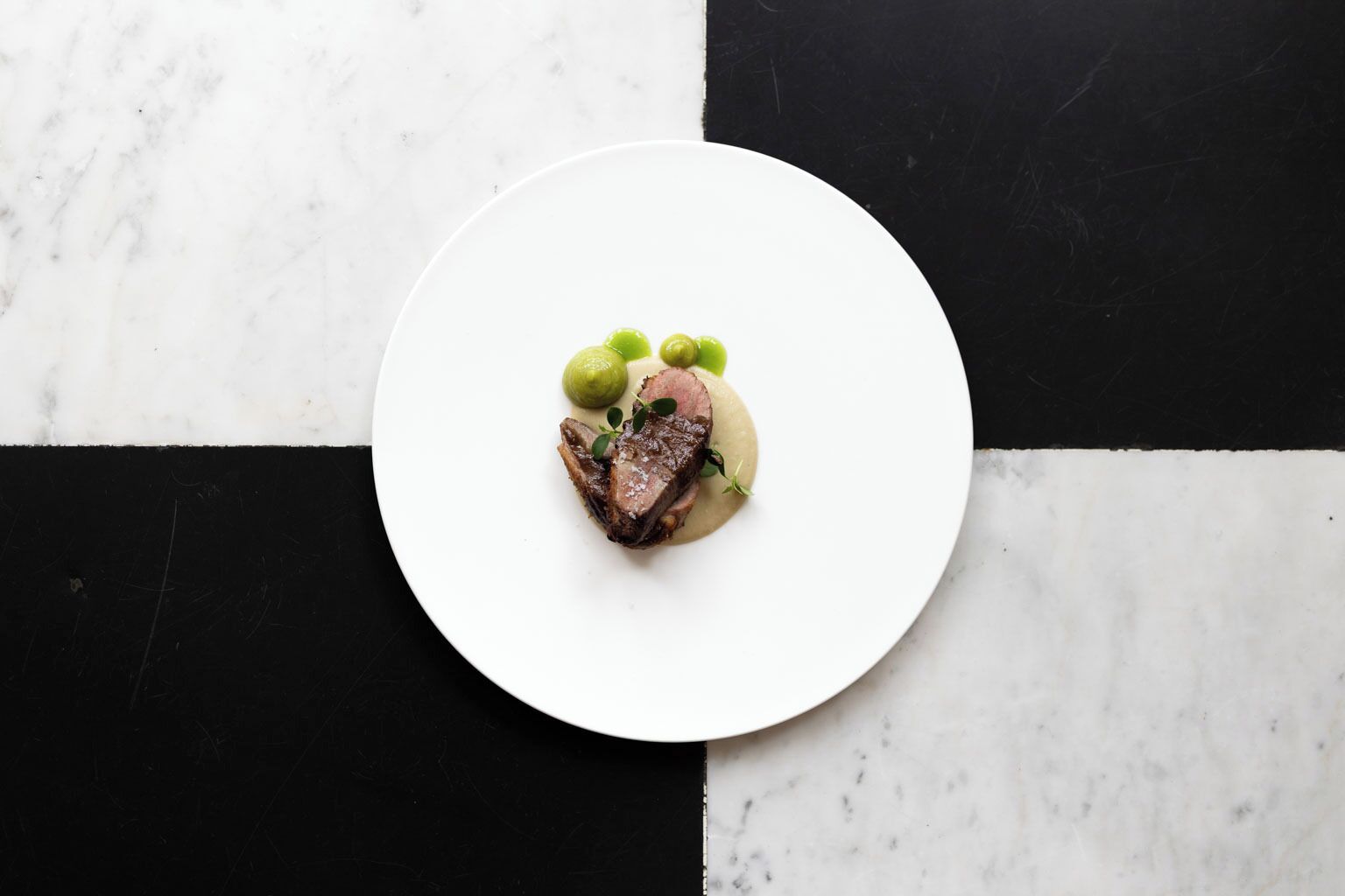 Une assiette blanche contenant un plat de viande, des olives vertes et une sauce crémeuse, le tout posé sur une surface en marbre noir et blanc.