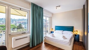 A hotel room with a large bed, a nightstand, a window with curtains, and a view of the city. - Rosabianca Boutique Hotel (Rapallo)