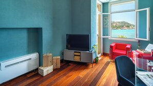 A room with teal walls, a wooden floor, a TV, a red chair, a window with a view of a coastal town, and a ceiling light. - Rosabianca Boutique Hotel (Rapallo)