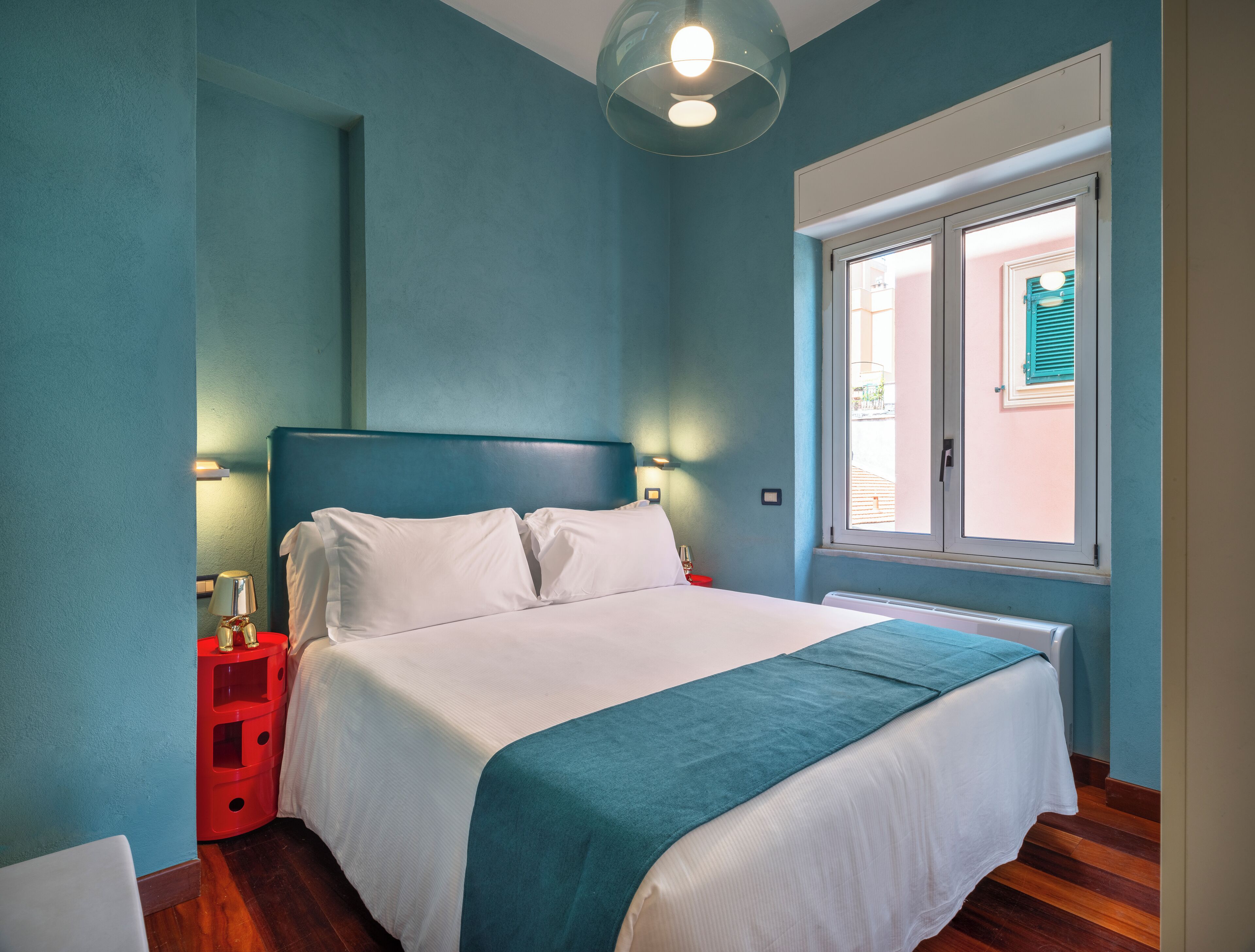 A bedroom with a bed, a red nightstand, a window, and a view of the outside.