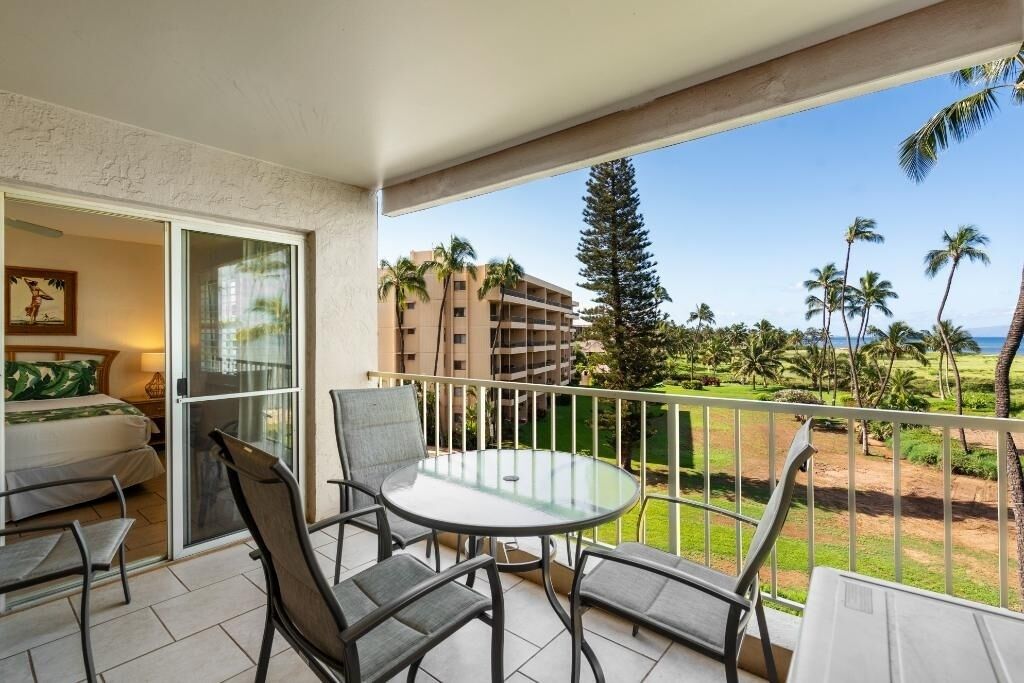 A balcony with a glass table and chairs, a bed with a green bedspread, and a view of a beach with palm trees and a building in the background.