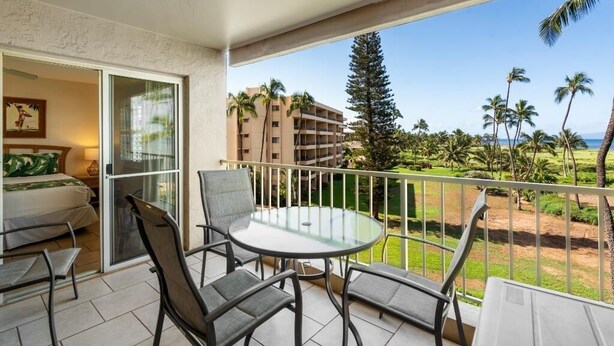 Ein Balkon mit Glastisch und Stühlen, ein Bett mit grüner Bettdecke und Blick auf einen Strand mit Palmen und einem Gebäude im Hintergrund.