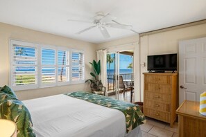 A hotel room with a bed, a television, a ceiling fan, a view of the ocean, and a balcony with chairs. - Menehune Shores #423 2 Bedroom Condo by RedAwning (Kihei)