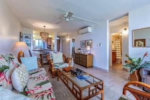 Un salon avec un canapé, une table basse et des fauteuils, un ventilateur de plafond, et un coin cuisine à l’arrière-plan.