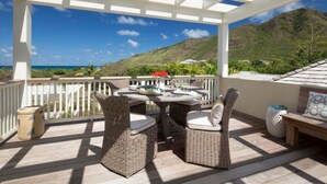 A covered outdoor area with wicker furniture, a table, and chairs, overlooking a mountainous landscape and the ocean.