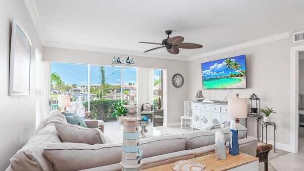 A living room with a large window, a ceiling fan, a TV, and a wooden table with decorative items. - Waterfront 2 Bed in Popular Olde Marco I Close to Snook Inn (Marco Island)