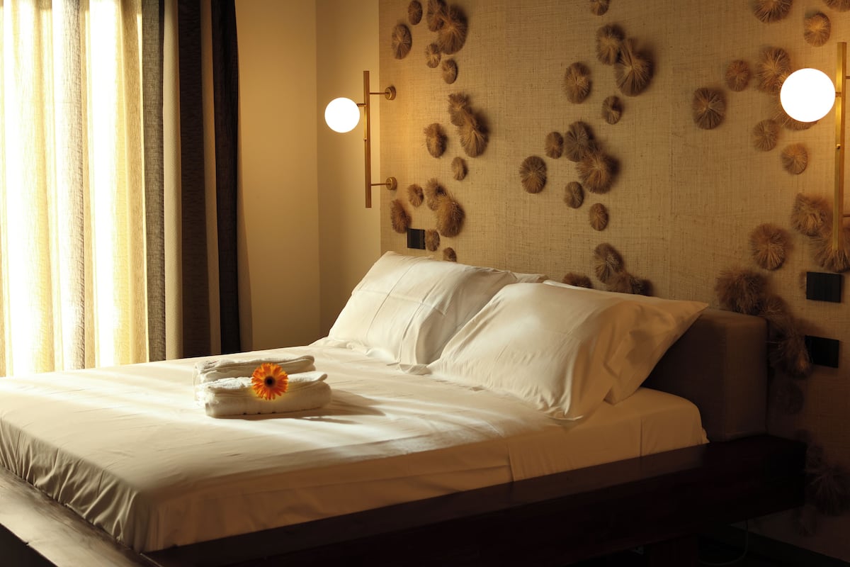 Un lit bien fait, avec du linge de lit blanc et une serviette ornée d’une fleur, un mur au motif texturé et deux lampes fixées au mur.