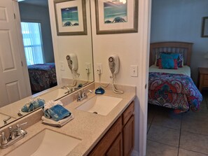 A bathroom with two sinks, a hairdryer, and a mirror. There is a bed with a colorful bedspread in the background.