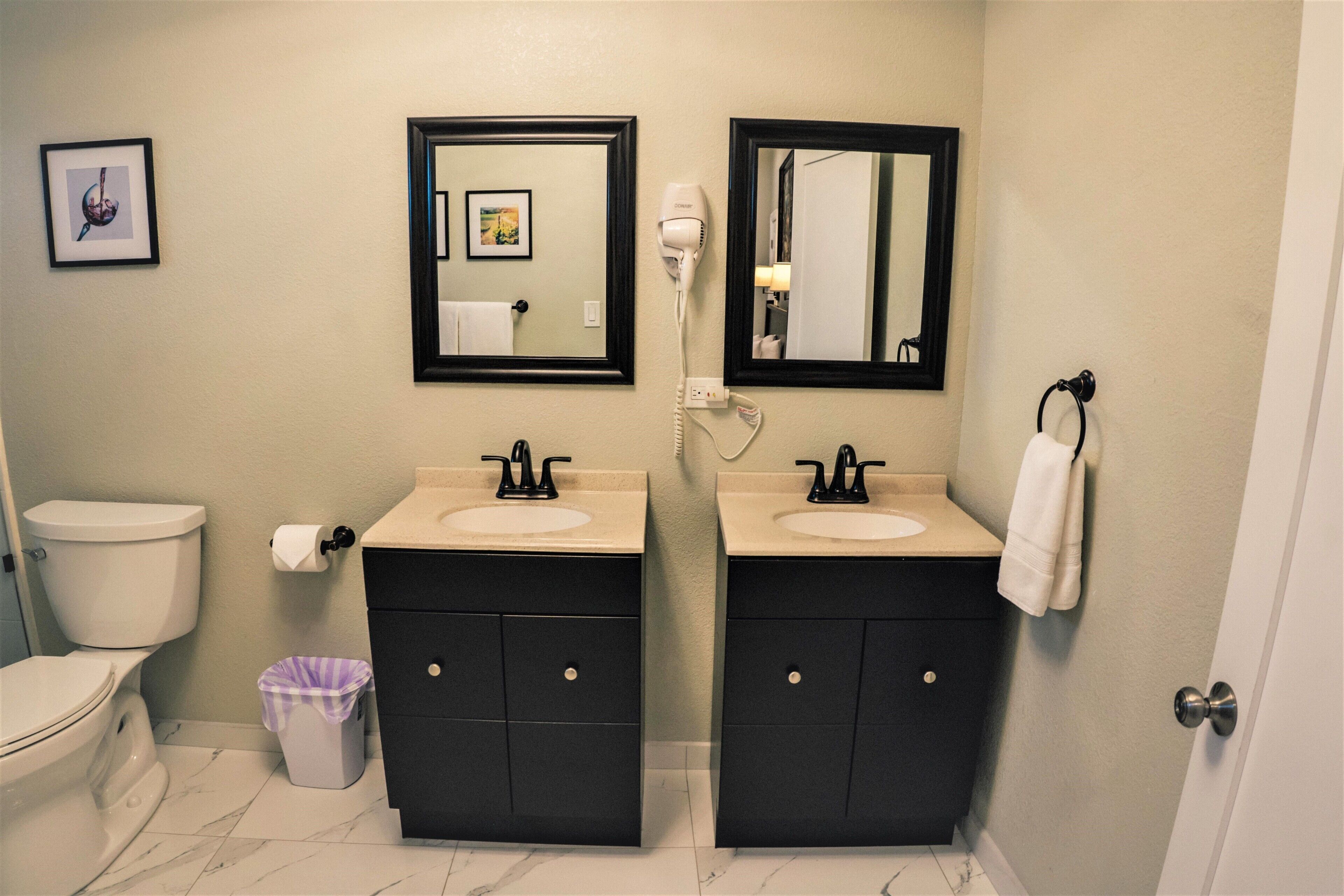 A bathroom with two sinks, a toilet, a trash bin, and a mirror.