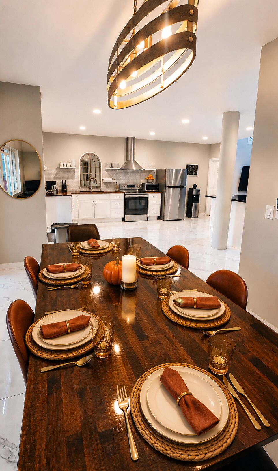 Une salle à manger moderne avec une longue table dressée pour six personnes, un lustre central et une cuisine en arrière-plan.