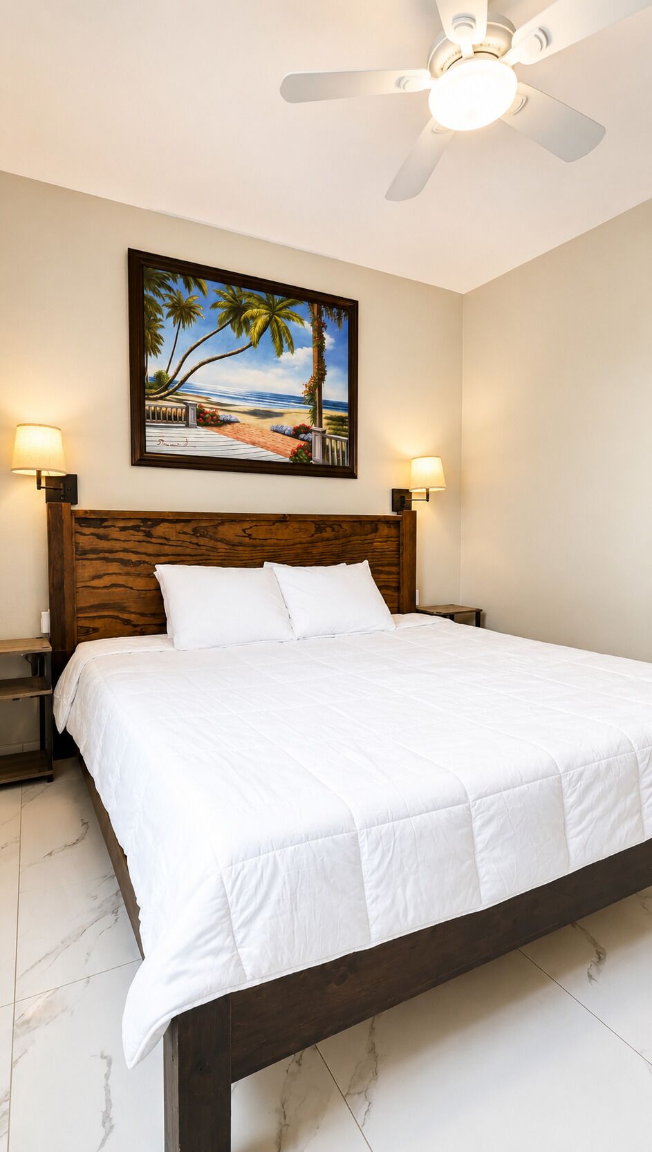 Un dormitorio con cabecera de madera, cama con ropa de cama blanca, ventilador de techo y un cuadro con motivos playeros en la pared.