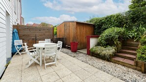 Eine Terrassenfläche mit einem runden Tisch und Stühlen, ein Holler Schuppen und eine kleine Treppe zu einem erhöhten Blumenbeet.