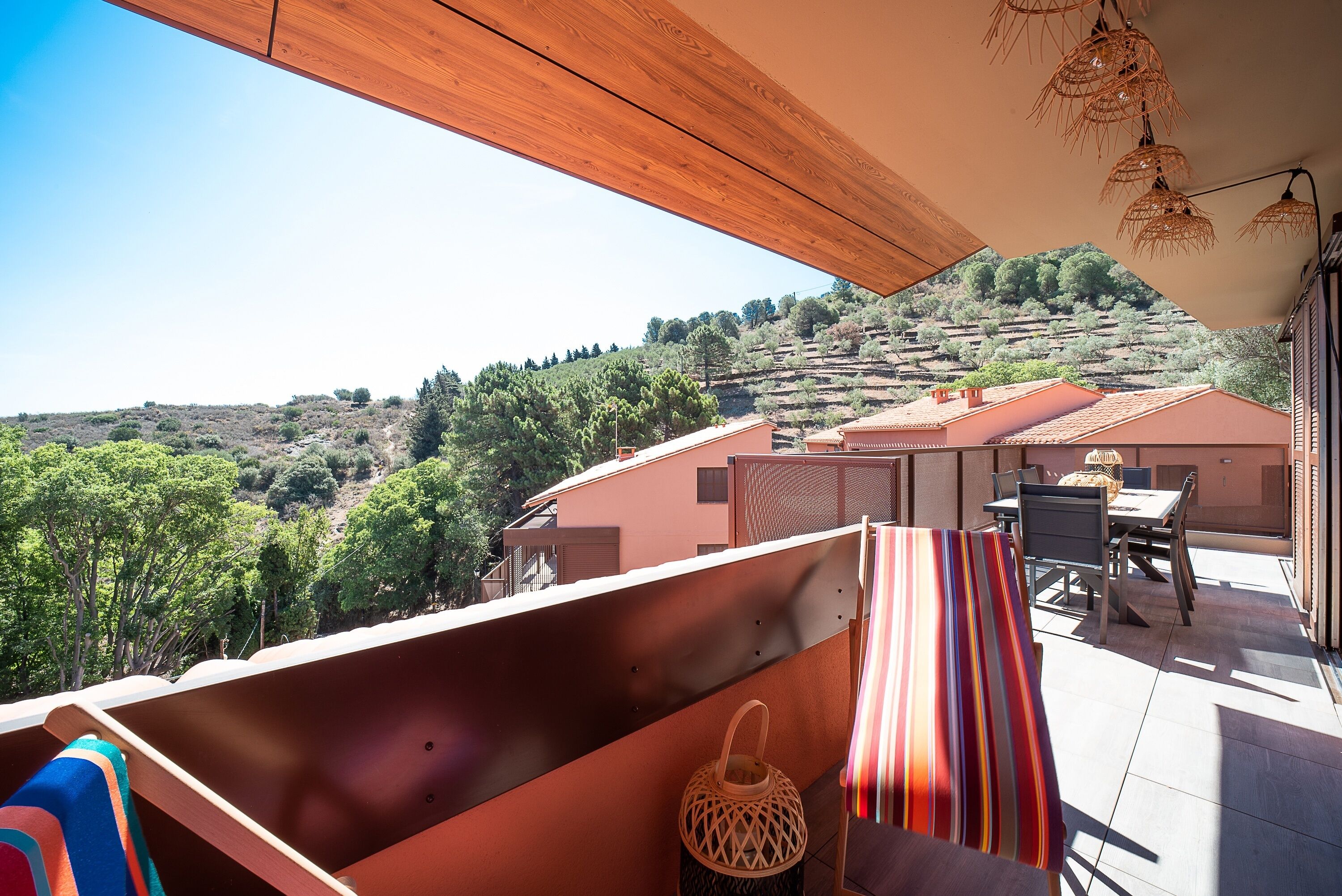 Ein Balkon mit Blick auf eine hügelige Landschaft, ein Tisch mit Stühlen und ein Korblampenlaterne.