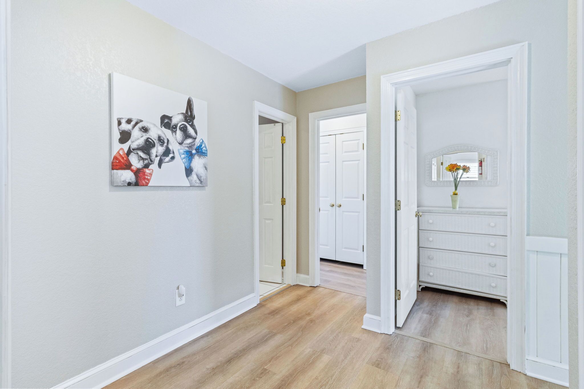 A hallway with light-colored walls and wooden flooring. There is a white door on the right and a white dresser with a vase of flowers.
