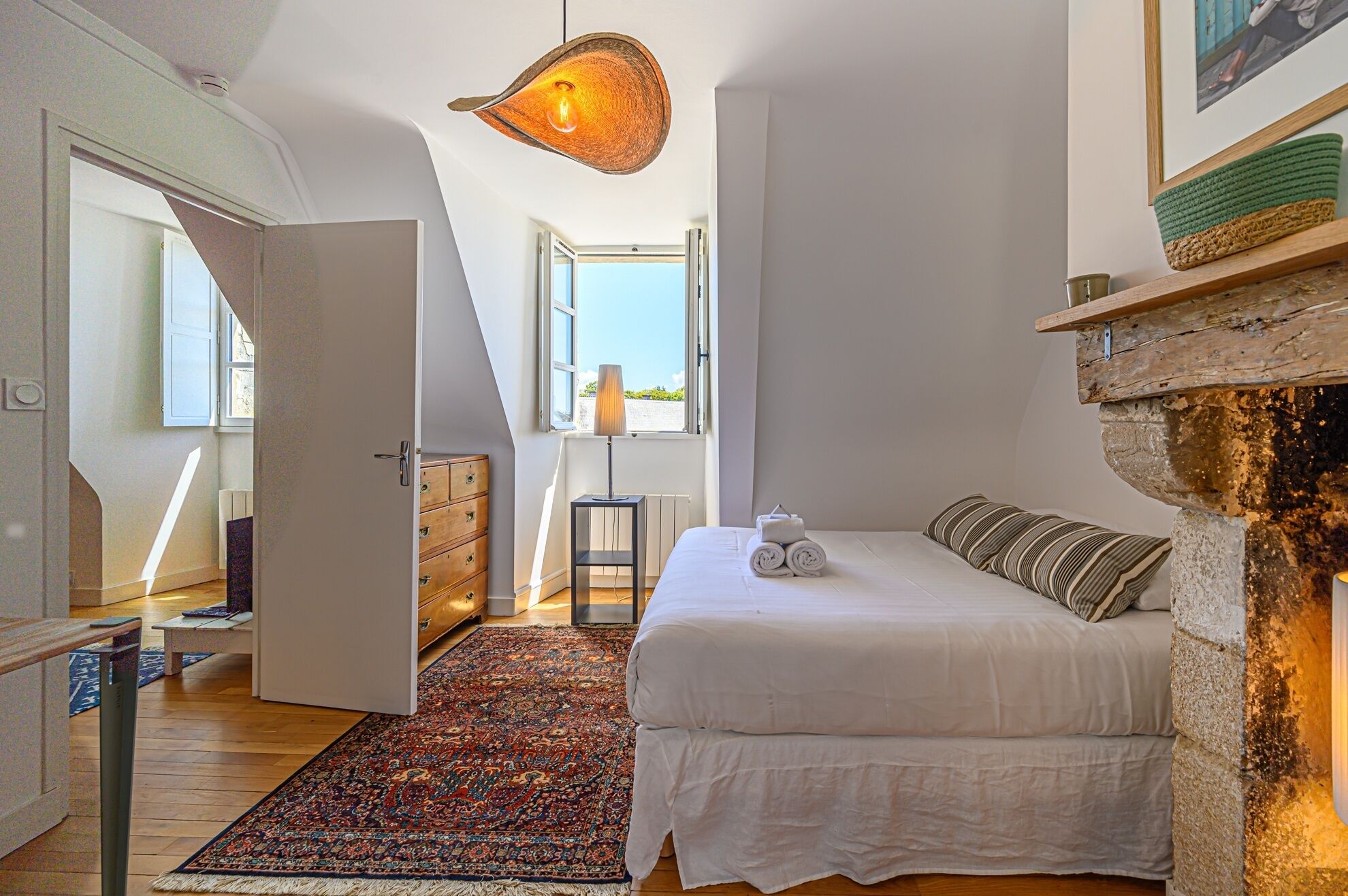 Une chambre avec un lit, un luminaire suspendu, une vue par la fenêtre et un mur en pierre.