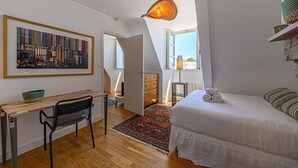 A bedroom with a bed, a desk, a chair, a lamp, a window, and a cityscape painting.