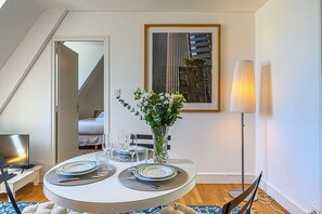 A dining area with a round table set for two, a vase with flowers, a framed cityscape picture, and a floor lamp.
