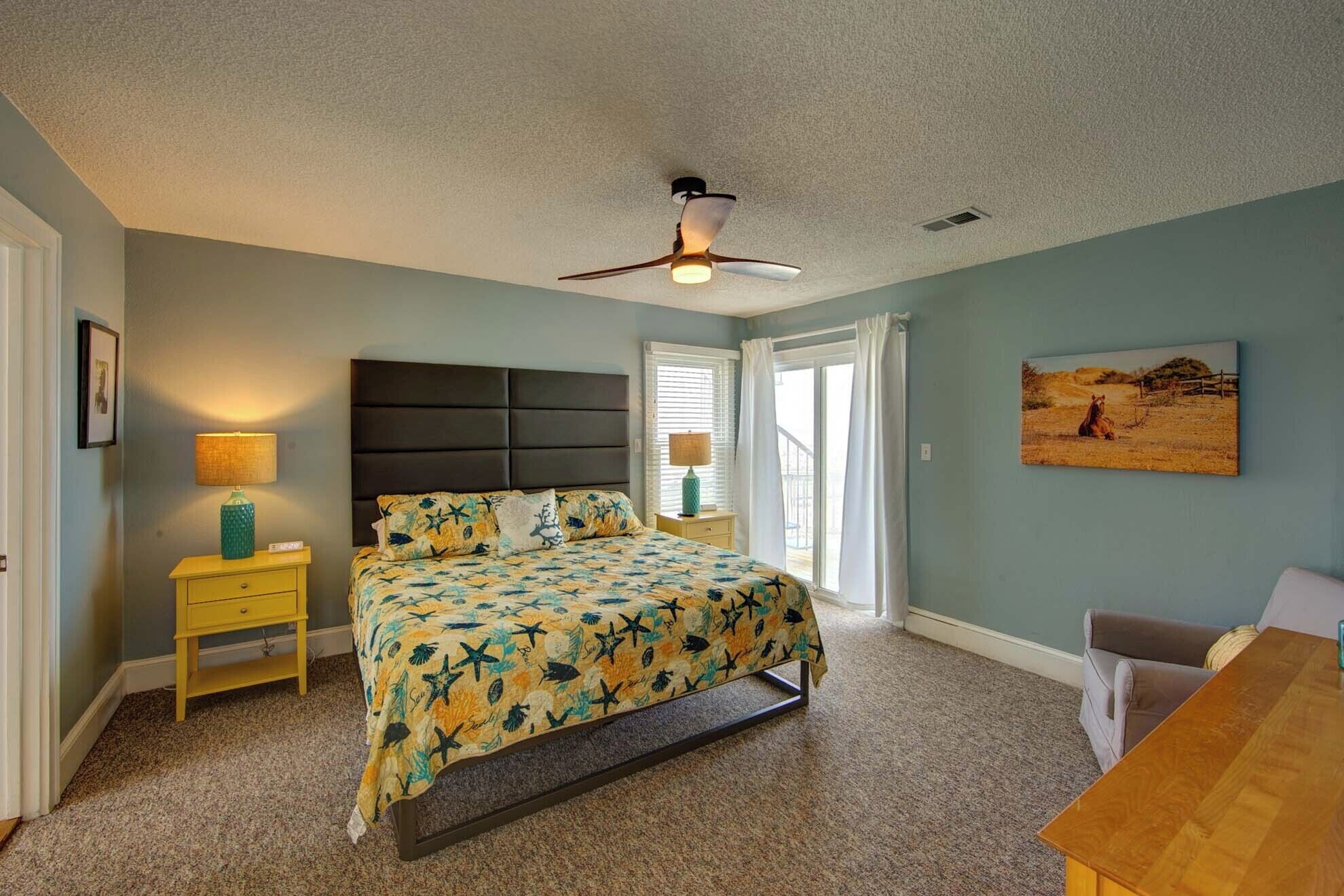 A bedroom with a bed, a nightstand, a desk, a ceiling fan, and a painting on the wall.