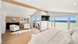 A spacious bedroom with a large bed, a view of the beach, and a modern kitchen in the background.