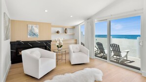 A modern living room with a fireplace, two white armchairs, a small table with a vase of flowers, and a view of the beach through large sliding glass doors.