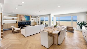 A modern living room with a fireplace, a dining area, and large windows overlooking a beach.
