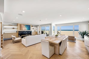 A modern living room with a fireplace, a dining area, and large windows overlooking a beach.