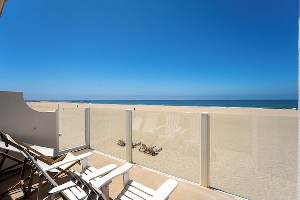 A beach view from a balcony with white chairs and a glass barrier.