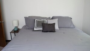 Una cama bien tendida con ropa de cama gris y varios almohadones, una mesita de noche pequeña con una lámpara, y una pared blanca lisa.