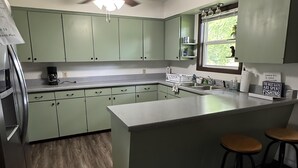 Una cocina con armarios verdes, una isla central con taburetes, un ventilador de techo y una ventana con vista a vegetación.