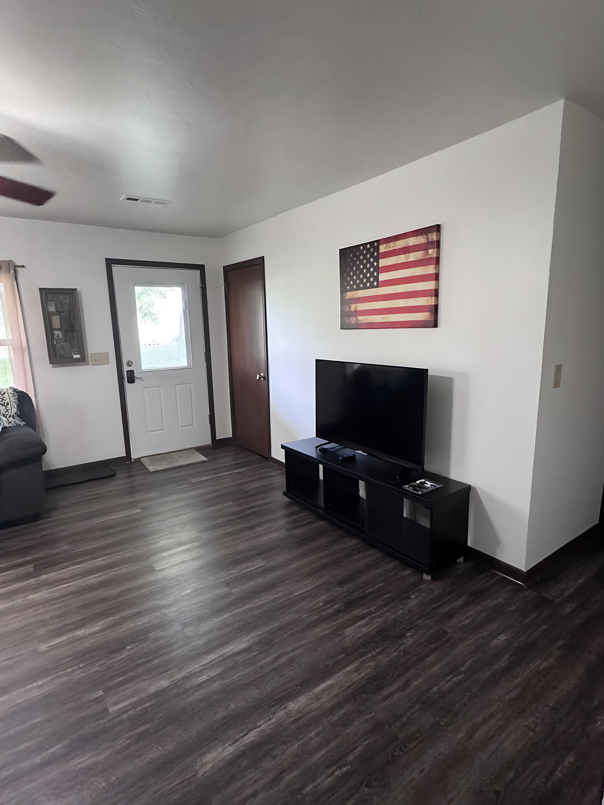 A living room with a dark wood floor, a flat-screen TV on a black stand, a door, and a large American flag on the wall.