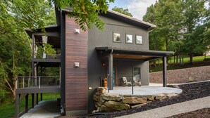 A modern two-story house with a covered patio, stone retaining wall, and a view of the surrounding wooded area. - Long Range Mtn Views Next to Jump Off Rock! Ultra Modern, Very Unique Open Floor (Hendersonville)