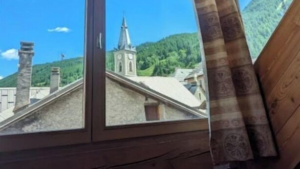 A wooden attic room with a large window overlooking a church and green hills. - Duplex 6/8 Pers 90 m² Le Bez Serre Chevalier 1400 (La Salle-les-Alpes)