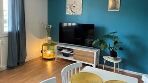 A living room with a teal wall, a flat-screen TV, a white cabinet, a small round table, and a potted plant. - Appartement : Le Petit Aubray (Châteauroux)