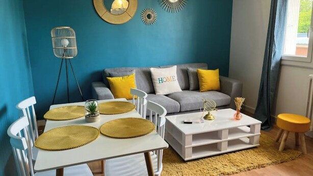A living room with a blue wall, a grey sofa with yellow cushions, a white coffee table, and a dining table with white chairs. - Appartement : Le Petit Aubray (Châteauroux)