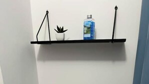 A small bathroom with a white toilet, a shelf above it holding a plant and a bottle, and a blue wall. - Appartement : Le Petit Aubray (Châteauroux)