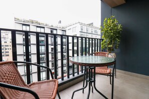 Un balcon agrémenté de fauteuils en osier et d’une table en verre, offrant une vue sur un paysage urbain avec des bâtiments modernes.