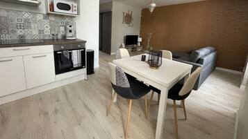 Una cocina moderna con armarios blancos, un microondas y una cafetera. Hay una mesa de comedor con dos sillas y un jarrón decorativo. Se ve un sofá gris y un televisor al fondo.