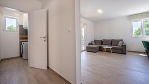 Un salon moderne avec un canapé d’angle, une petite table ronde et un fauteuil vert. Une porte donne accès à une buanderie équipée d’un lave-linge.