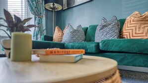 A green sofa with patterned pillows, a small round table with a book, and a potted plant. - 3BR Chic House withParking Sleeps 8, Big Groups! (Portsmouth)