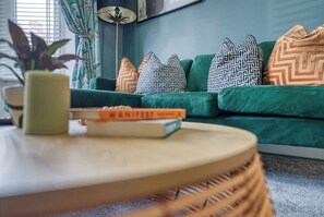 Ein grรผnes Sofa mit gemusterten Kissen, ein kleiner runder Tisch mit einem Buch und ein Topfpflanze.