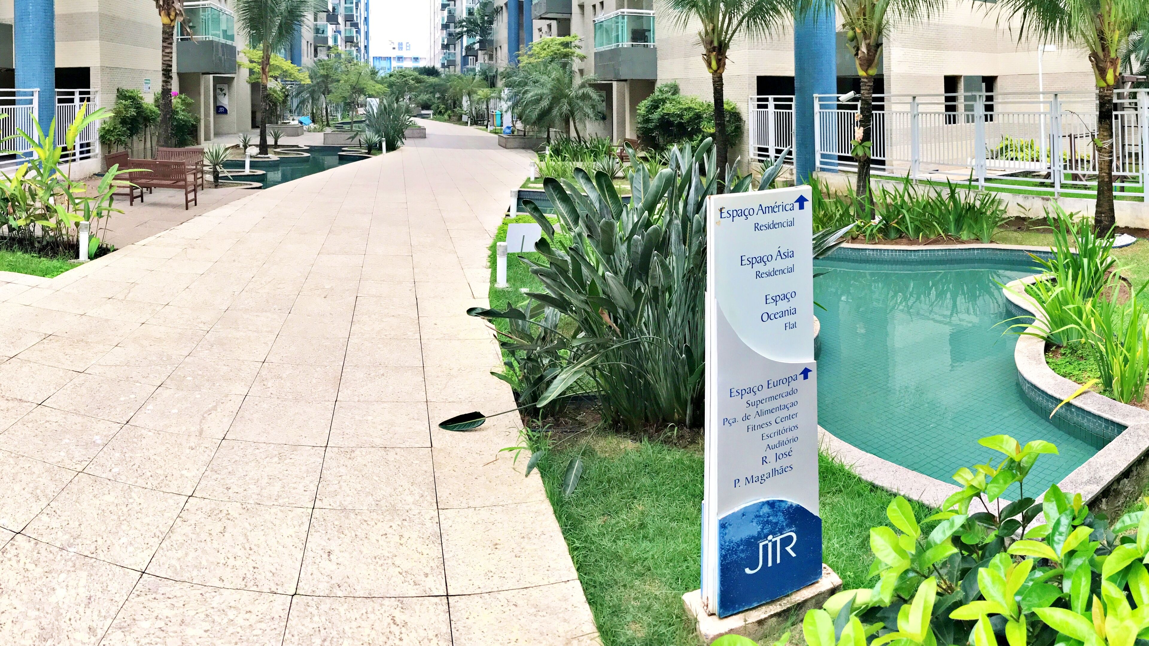 A residential area with a paved walkway, a sign indicating various amenities like a fitness center and auditorium, and a pool area.