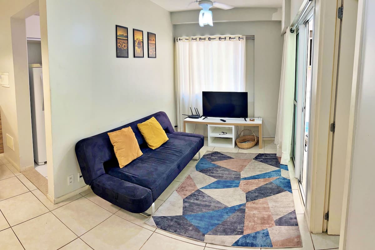 Un salon avec un canapé bleu foncé, deux coussins jaunes, un téléviseur à écran plat posé sur un meuble en bois, un tapis à motifs et un ventilateur de plafond.