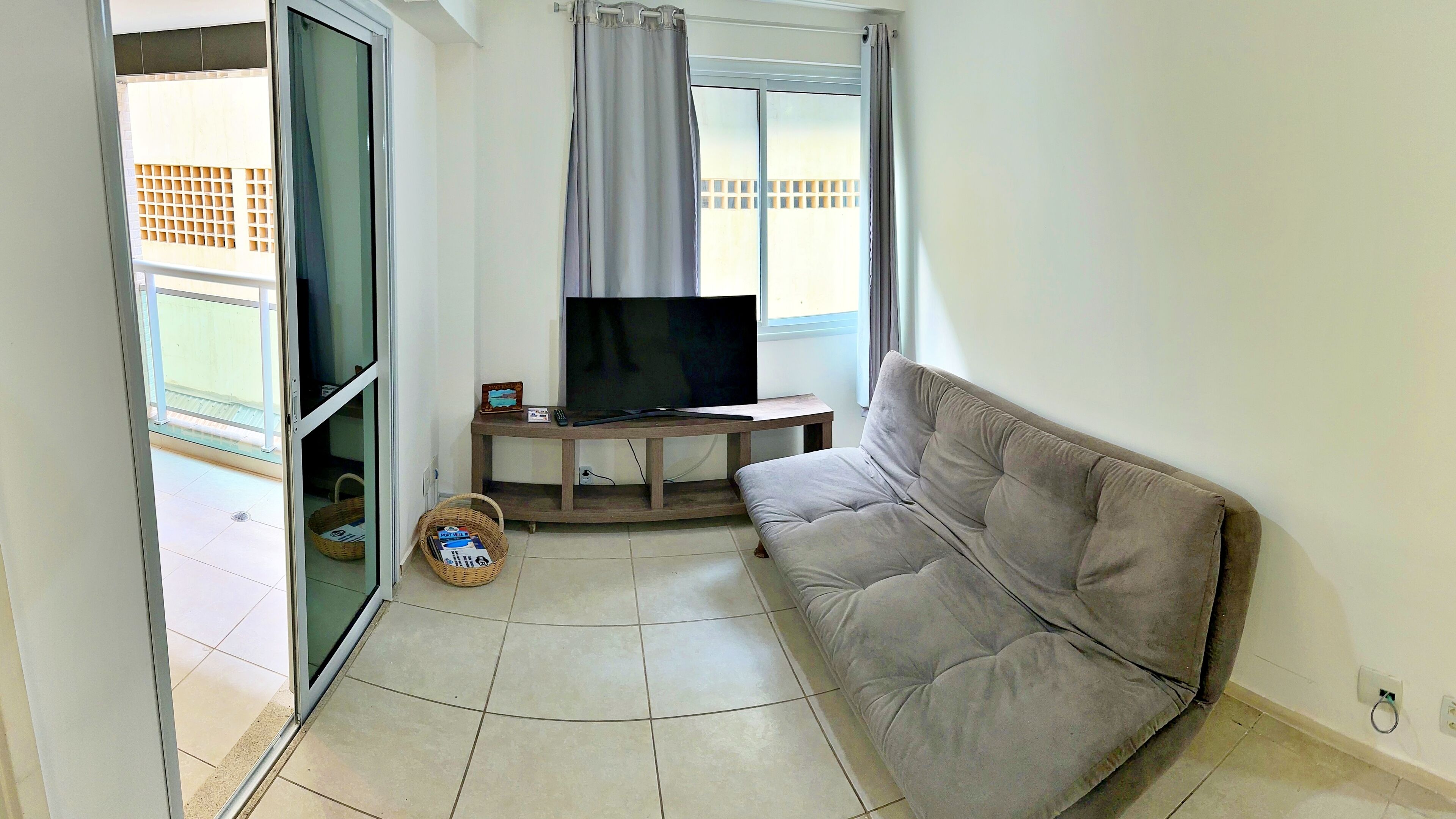 A living room with a grey sofa, a flat-screen TV on a wooden stand, a wicker basket, and a sliding glass door leading to an outdoor area.