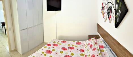 Un dormitorio con una cama cubierta de ropa de cama floral, un armario blanco y un televisor montado en la pared.