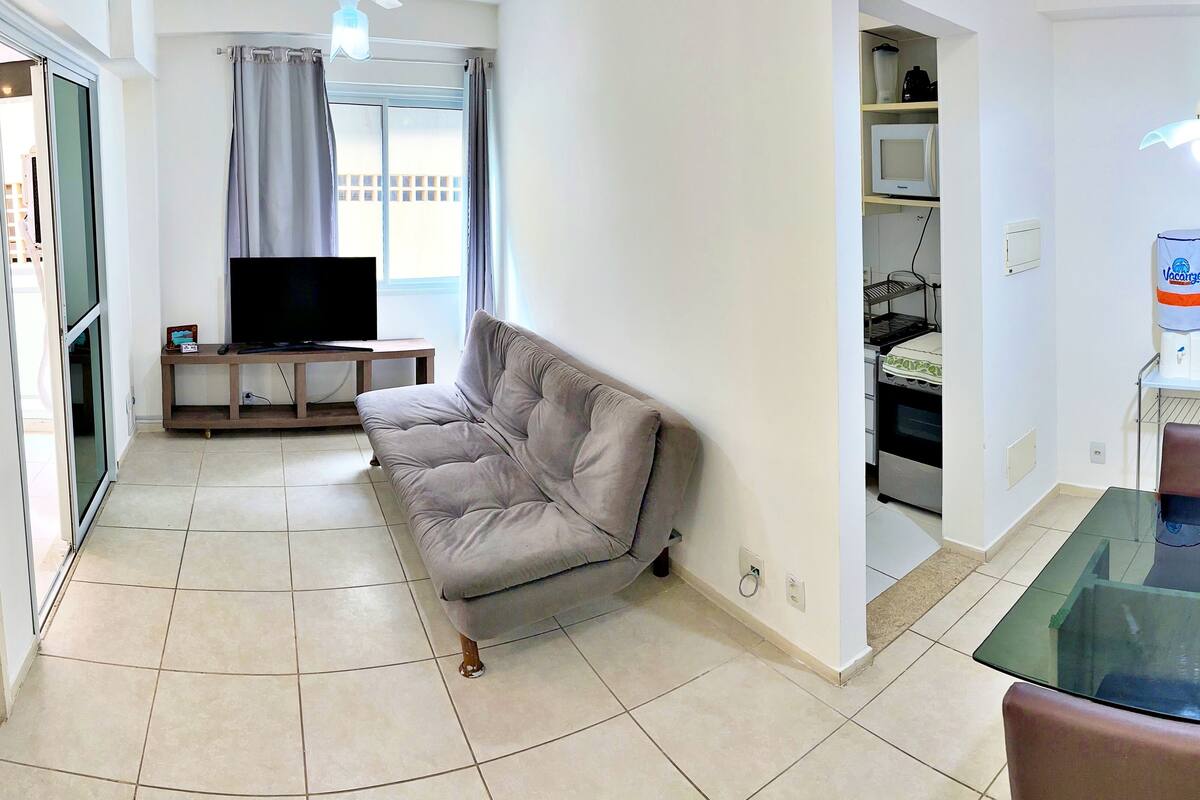 Un salon avec un canapé gris, un téléviseur posé sur un meuble en bois, et un coin cuisine équipé d’un four à micro-ondes et d’un four traditionnel.