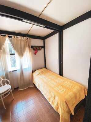 Pequeña habitación con cama individual, una silla, una ventana con cortinas y un gancho en la pared con una bolsa.
